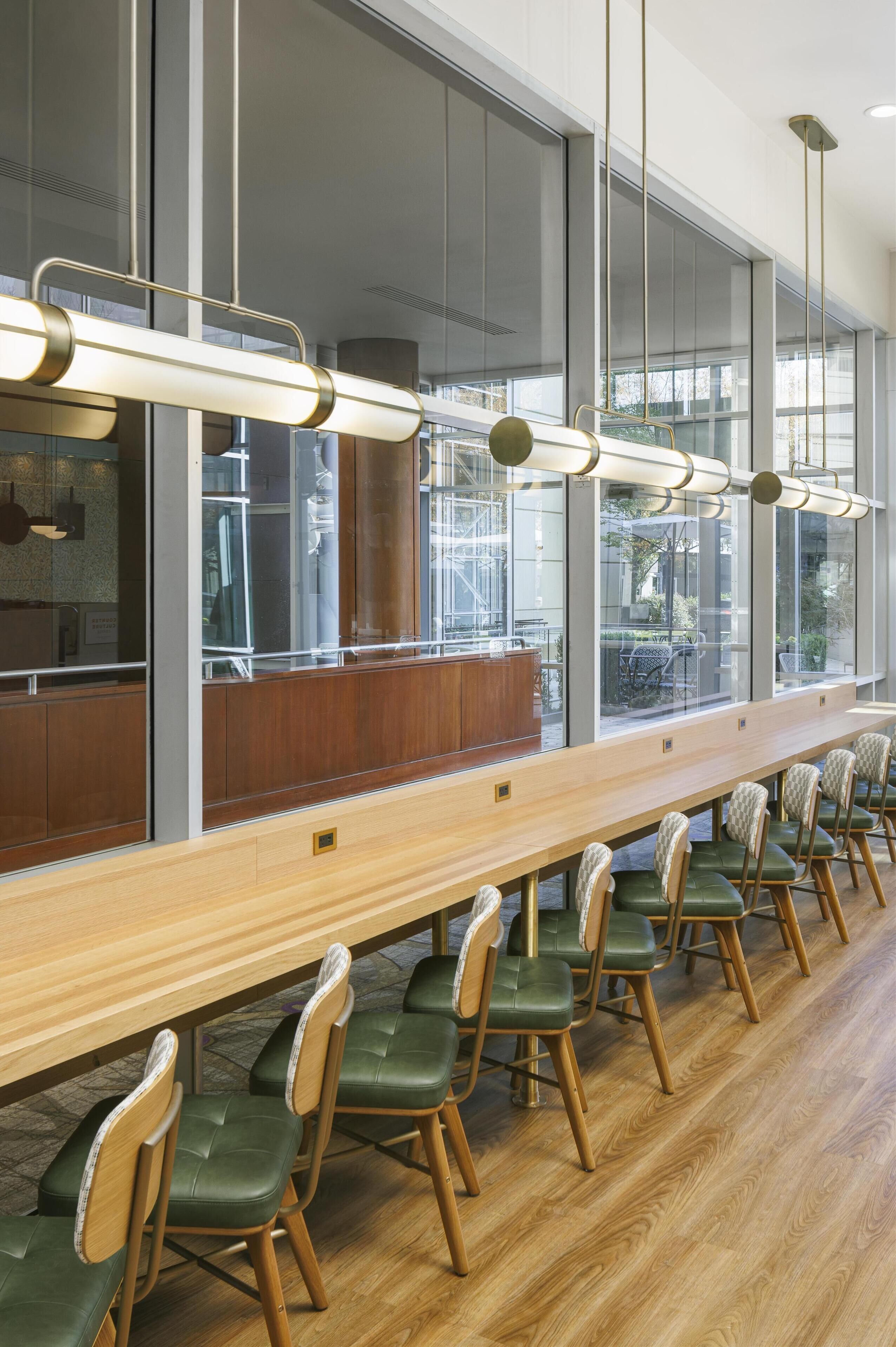 A long wooden table with green upholstered chairs in a modern interior with large windows and pendant lighting.