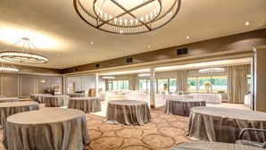 Una amplia sala de conferencias con varias mesas redondas cubiertas con manteles grises, ventanas grandes con cortinas y una lámpara de araña central.