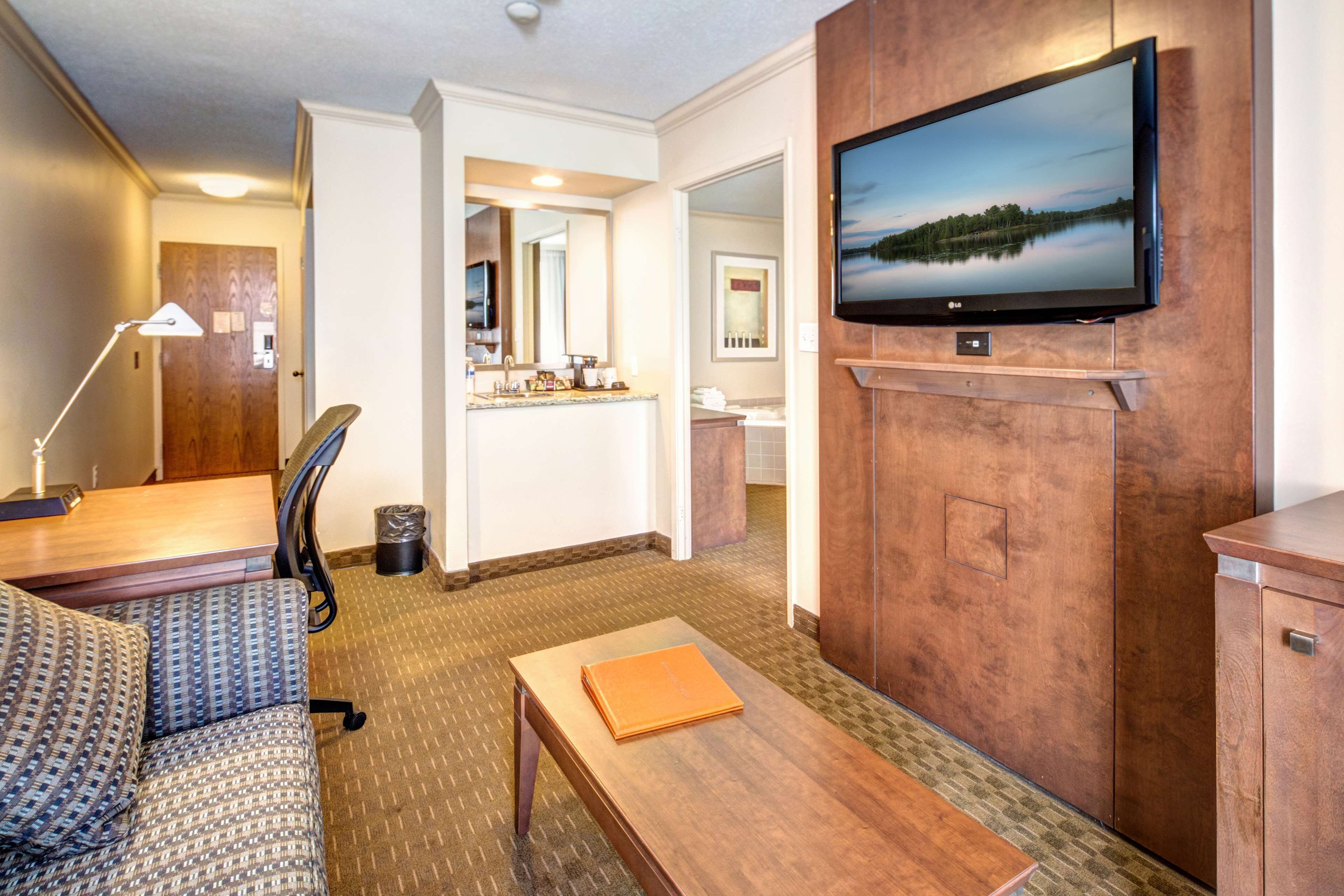 A hotel room with a sofa, a wooden table, a TV mounted on the wall, and a bathroom visible through an open door.