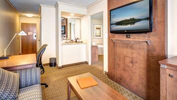 A hotel room with a sofa, a wooden table, a TV mounted on the wall, and a bathroom visible through an open door.