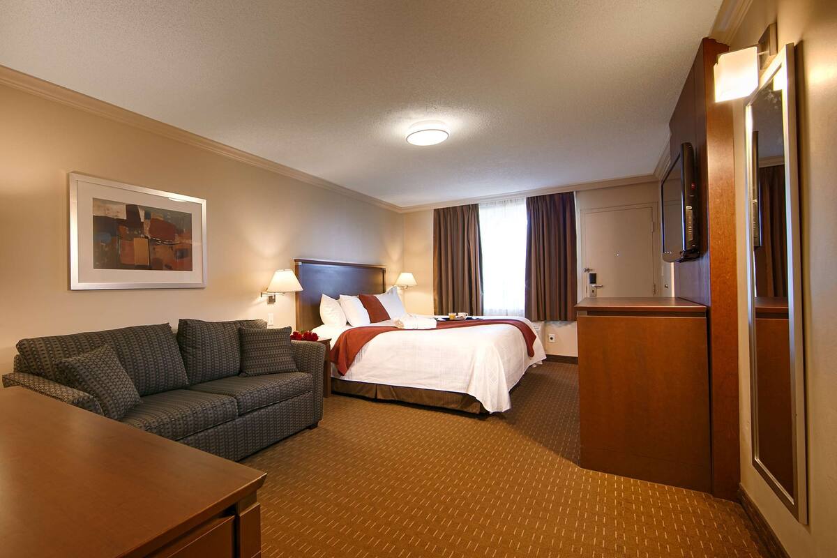 A hotel room with a bed, a sofa, a desk, a TV, and a painting on the wall.