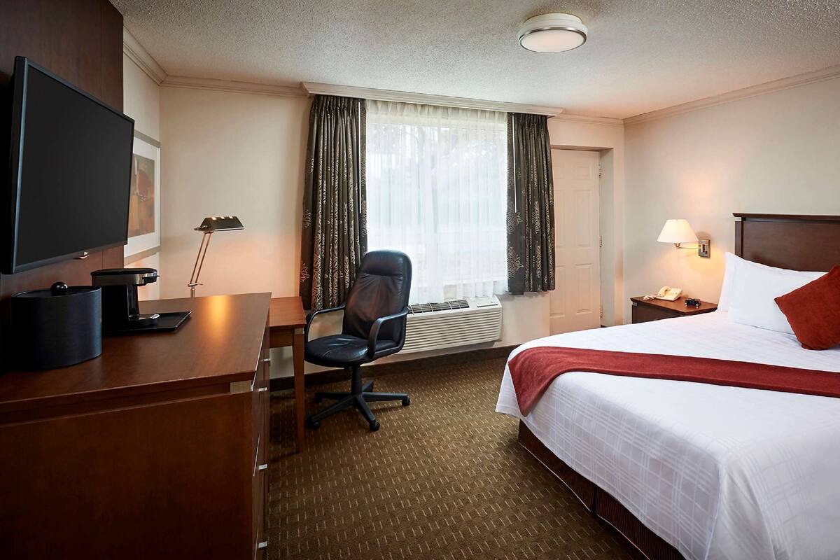 A hotel room with a bed, a desk with a TV, a chair, a lamp, and an air conditioning unit.