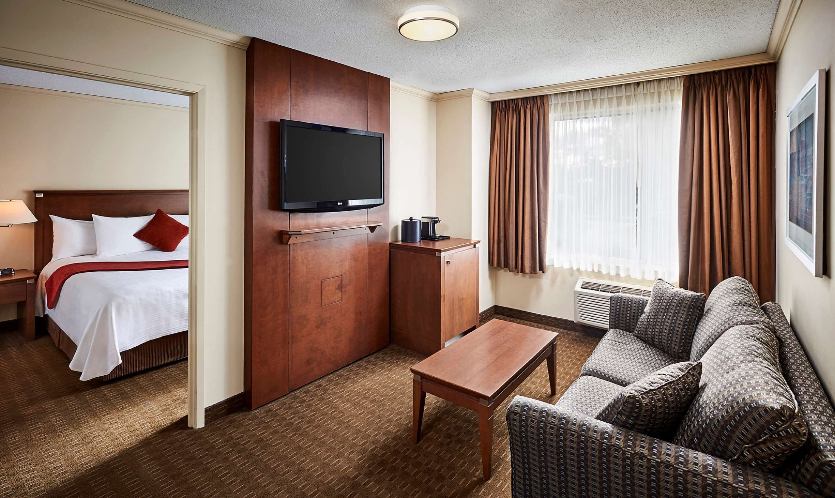 A hotel room with a bed, a TV mounted on a wooden panel, a small table, a sofa, and a window with curtains.