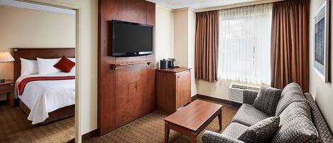 A hotel room with a bed, a TV mounted on a wooden panel, a small table, a sofa, and a window with curtains.