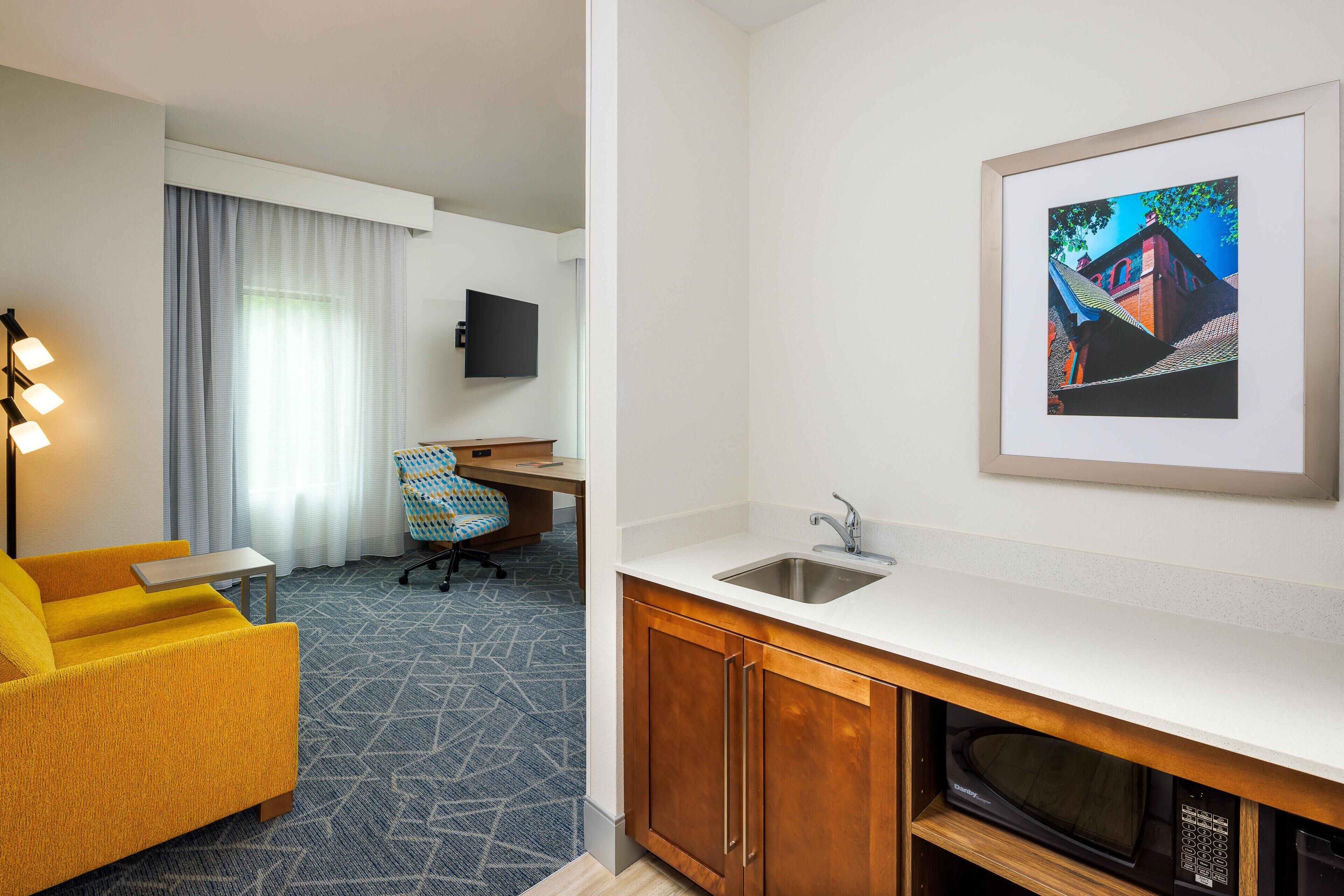 A hotel room with a kitchenette, a sofa, a desk, a chair, and a TV.