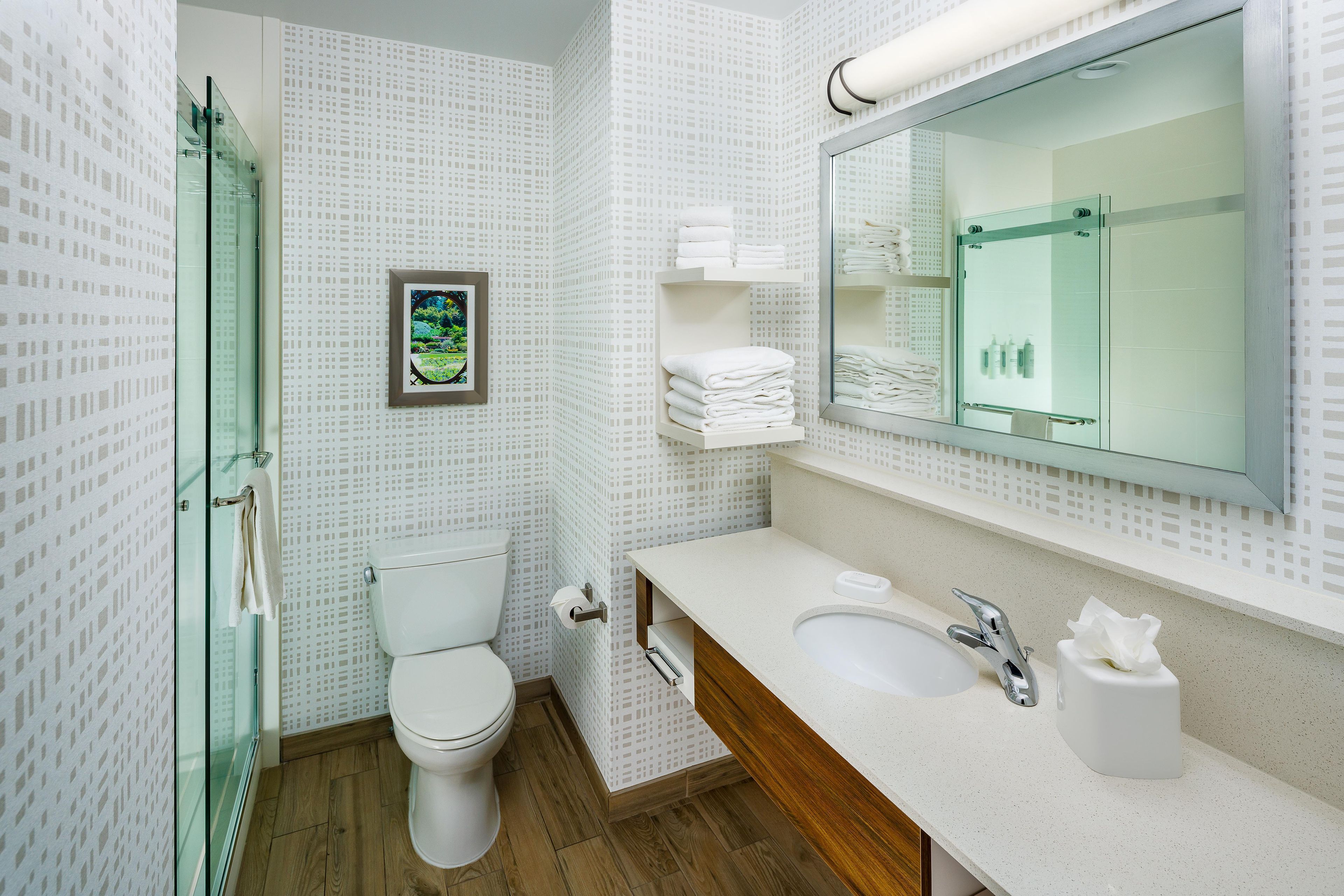 Ein modernes Badezimmer mit Toilette, einer Duschkabine aus Glas, einem Waschbecken mit Spiegel und ordentlich gefalteten Handtüchern auf einem Regal.