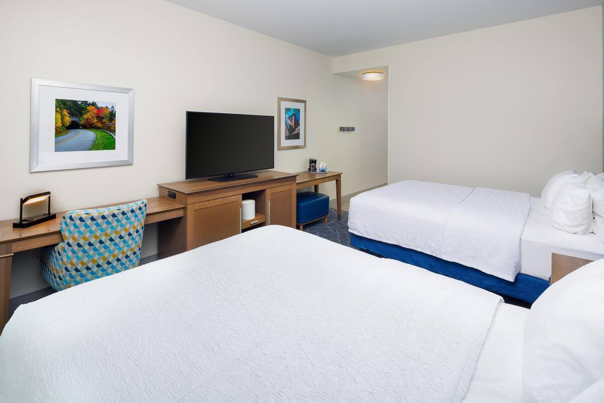 A hotel room with two beds, a desk with a television, and framed pictures on the wall.