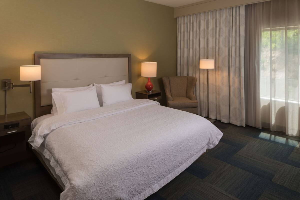 Een hotelkamer met een groot bed, twee bedlampen, een stoel en een raam met gordijnen.