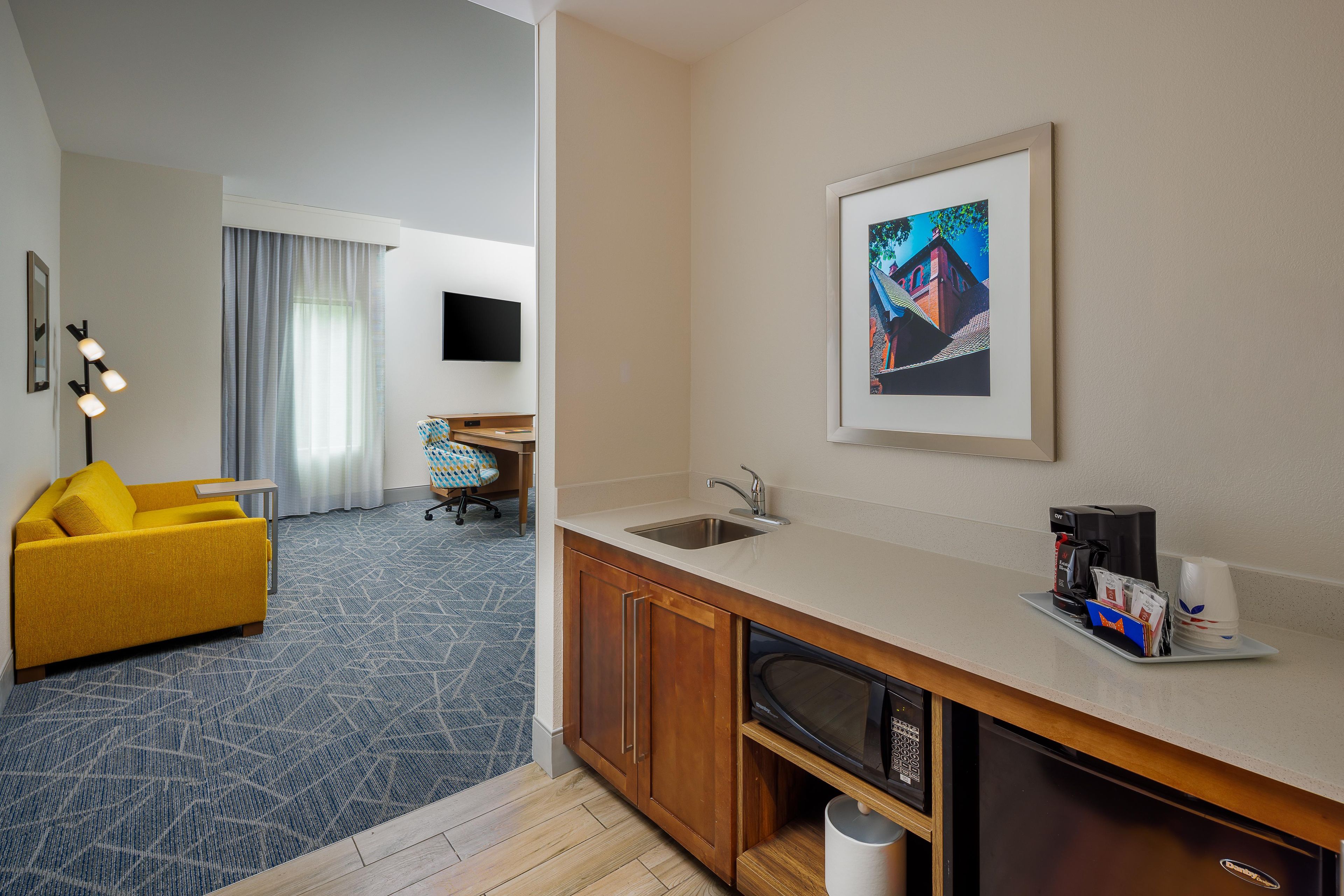 A hotel room with a kitchenette, a yellow sofa, a flat-screen TV, and a framed picture on the wall.