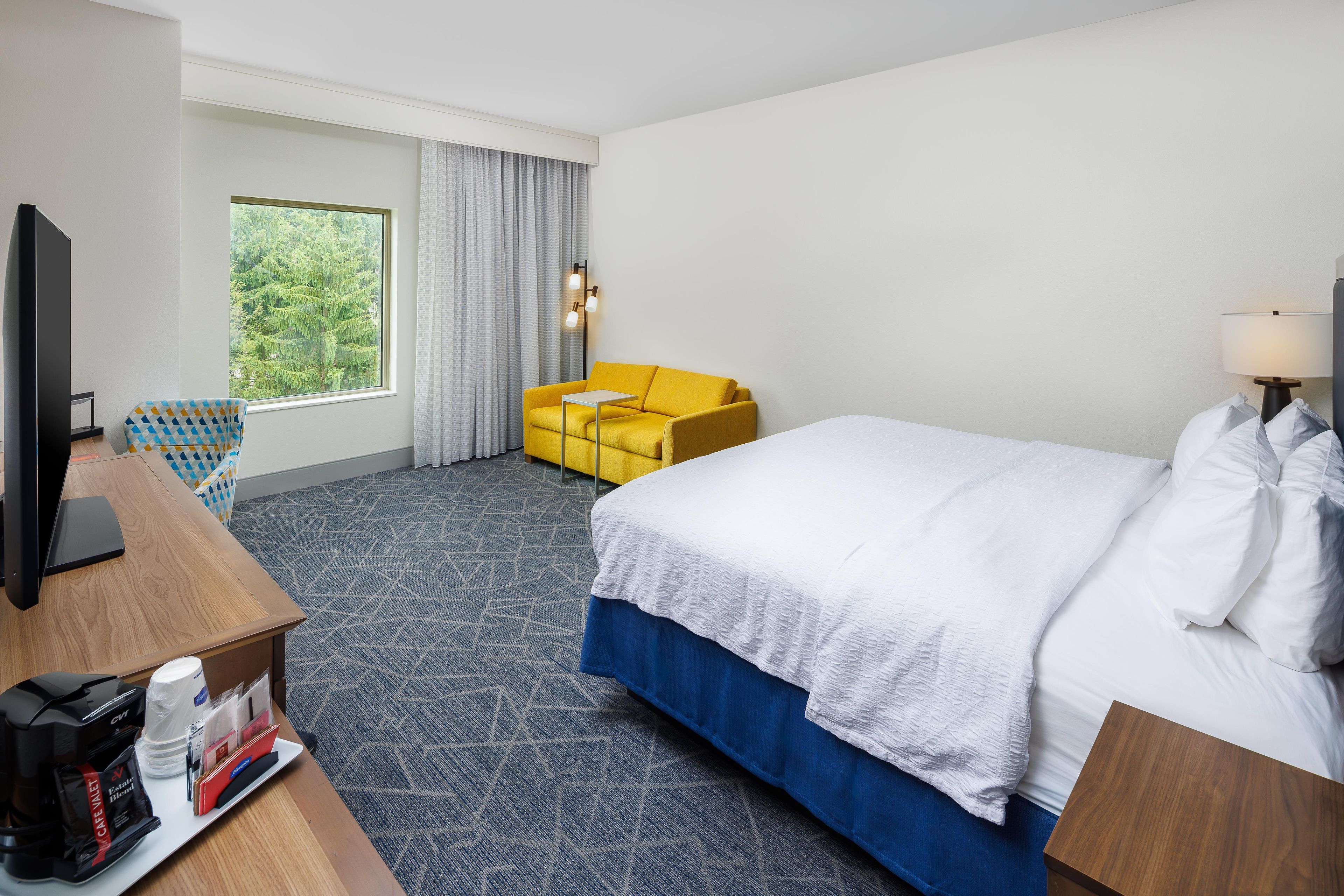 A hotel room with a bed, a TV, a desk with a coffee maker, a chair, and a window with curtains.