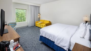 A hotel room with a bed, a TV, a desk with a coffee maker, a chair, and a window with curtains.