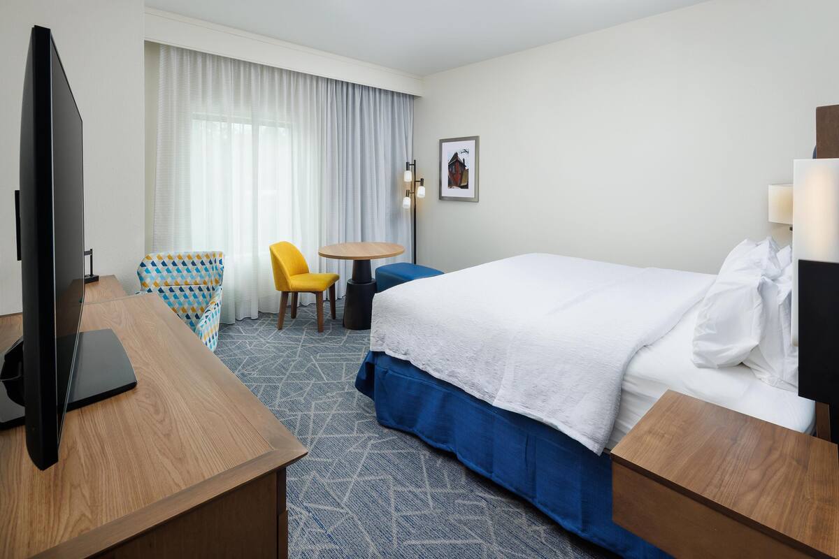 Een hotelkamer met een bed, een bureau met een televisie, een stoel, een kleine tafel en een schilderij aan de muur.