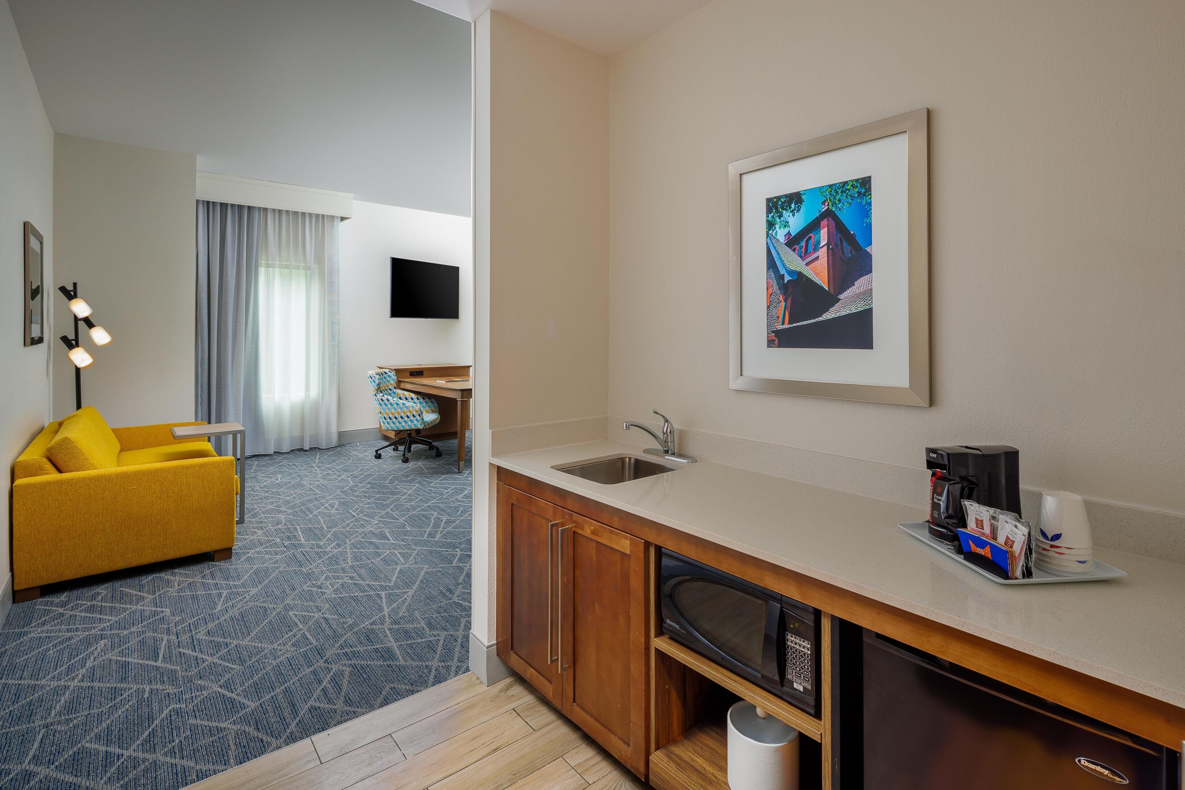 Quarto de hotel com cozinha compacta, sofá amarelo, televisão de tela plana e um quadro na parede.