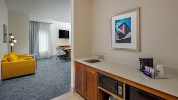 Quarto de hotel com cozinha compacta, sofá amarelo, televisão de tela plana e um quadro na parede.