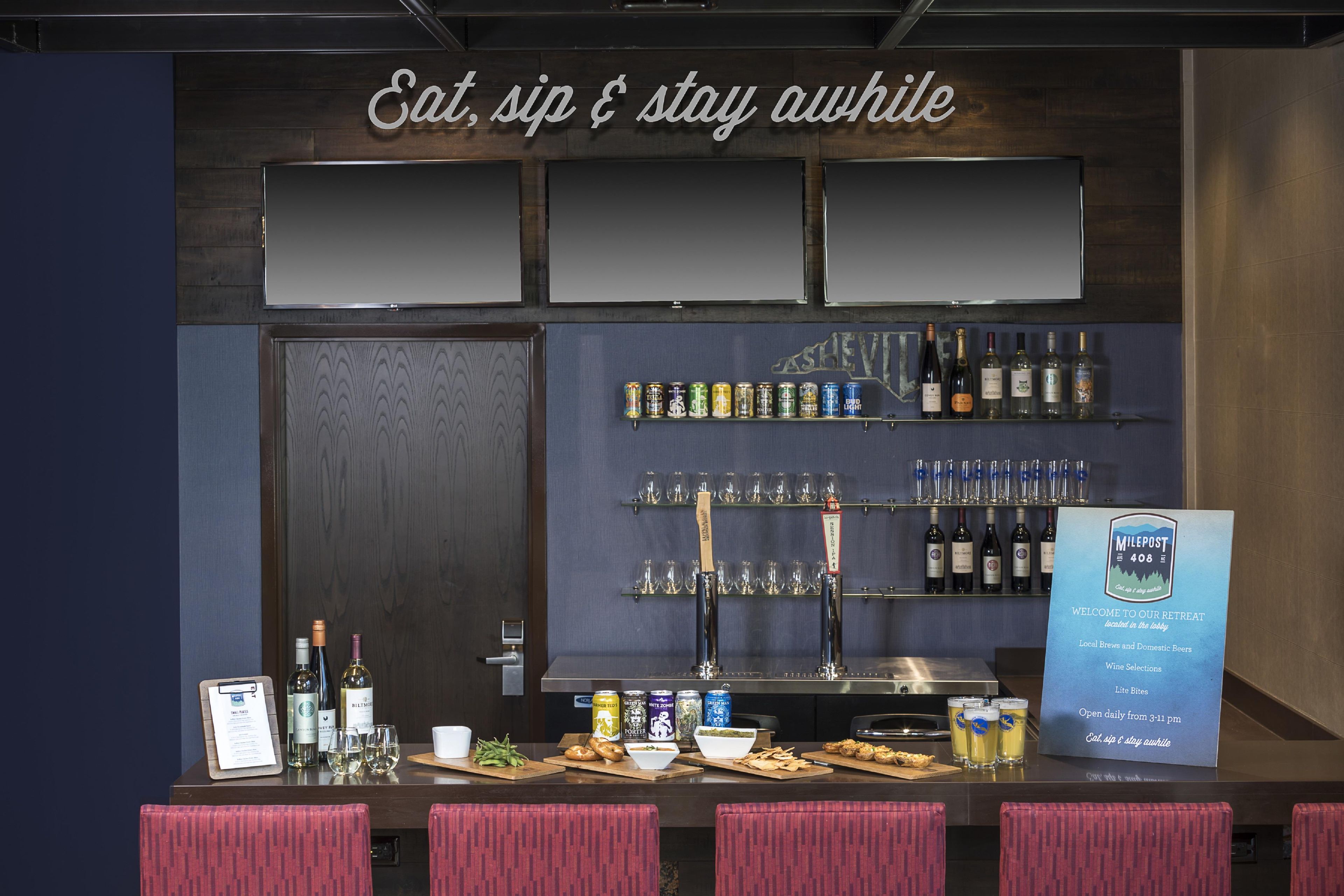 A bar area with a sign that reads 'Eat, sip & stay awhile.' There are shelves with bottles and cans, a counter with glasses and snacks, and a menu board.