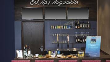A bar area with a sign that reads 'Eat, sip & stay awhile.' There are shelves with bottles and cans, a counter with glasses and snacks, and a menu board.