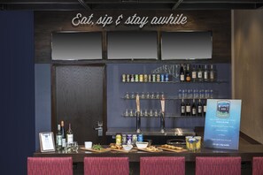 A bar area with a sign that reads 'Eat, sip & stay awhile.' There are shelves with bottles and cans, a counter with glasses and snacks, and a menu board.