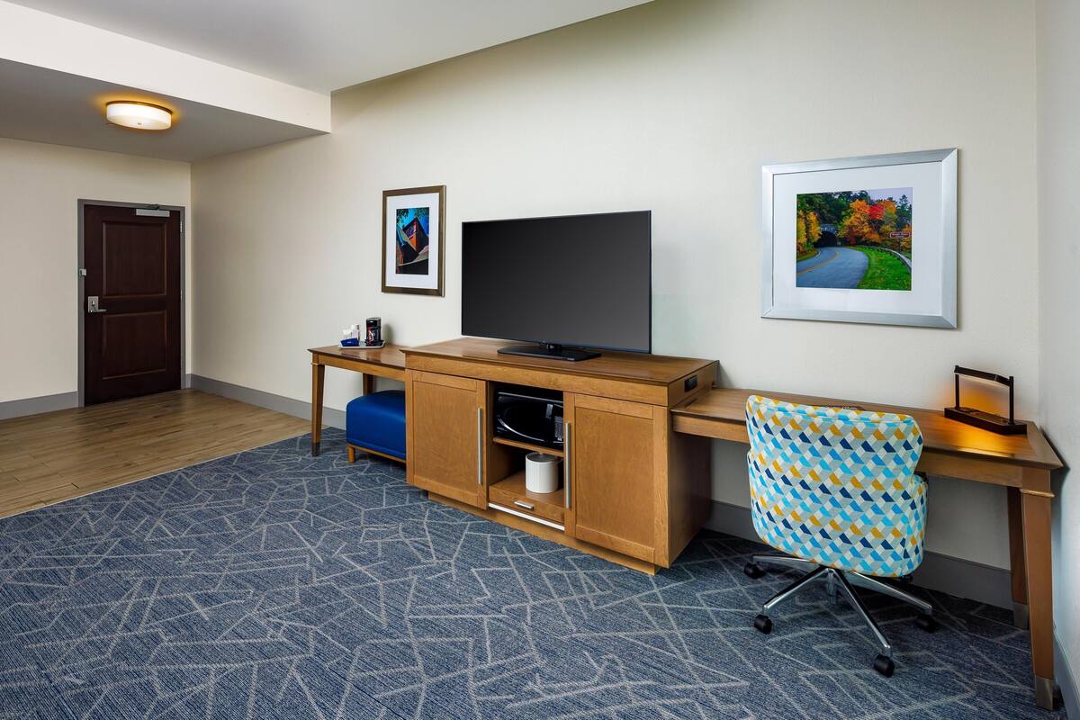 A hotel room with a wooden desk, a television, a framed picture, a patterned chair, and a blue carpet.