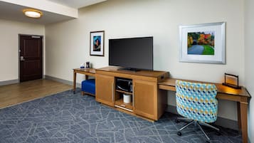 Quarto de hotel com escrivaninha de madeira, televisão, quadro emoldurado, cadeira estampada e tapete azul.