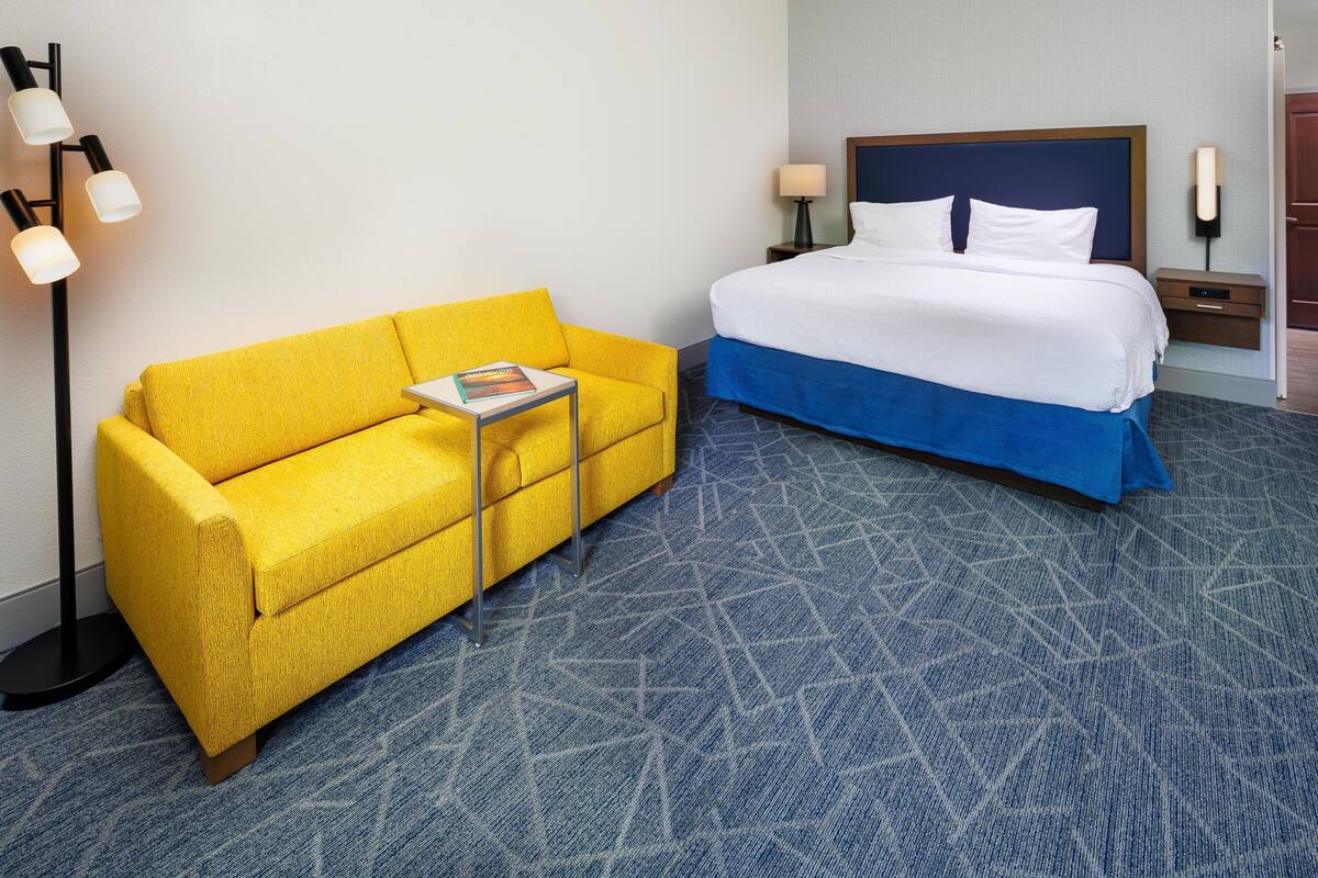 Een hotelkamer met een gele bank, een bed met een blauwe deken, een kamerbreed tapijt en een vloerlamp.