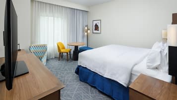 A hotel room with a bed, a desk with a TV, a chair, a small table, and a picture on the wall.