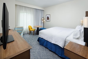 A hotel room with a bed, a desk with a TV, a chair, a small table, and a picture on the wall.