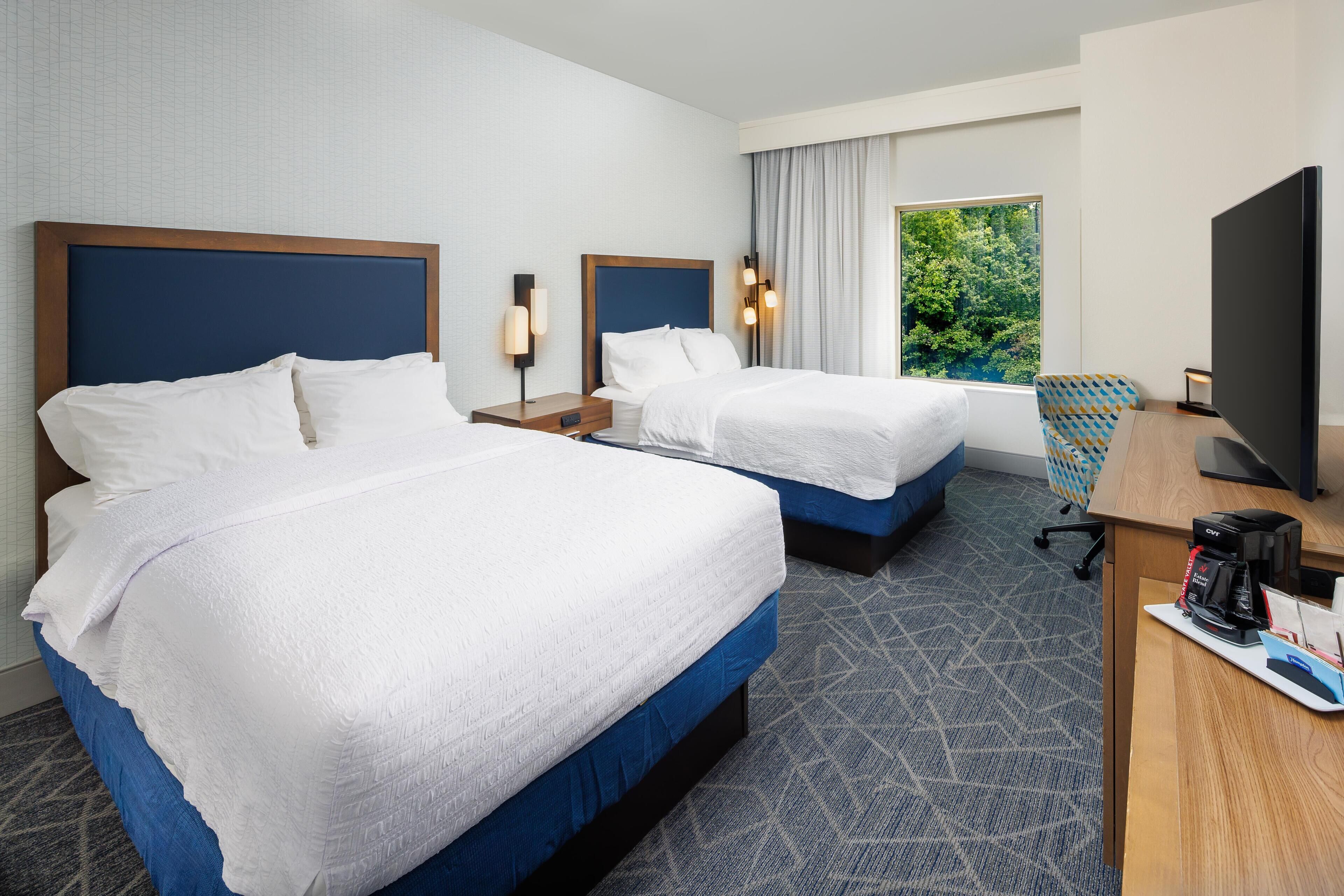 A hotel room with two beds, a desk with a coffee maker, a TV, and a window with a view of trees.