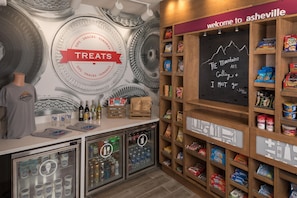 A snack bar with a 'TREATS' sign, shelves stocked with snacks, a chalkboard with a quote, and a refrigerator with beverages.