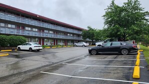 Un motel de varios pisos con techo rojo y balcones. Se ven varios coches aparcados en el estacionamiento, y un árbol es visible a la derecha.