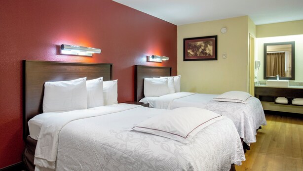 Habitación de hotel con dos camas, una pared roja, un cuadro enmarcado y un baño visible a través de una puerta abierta.