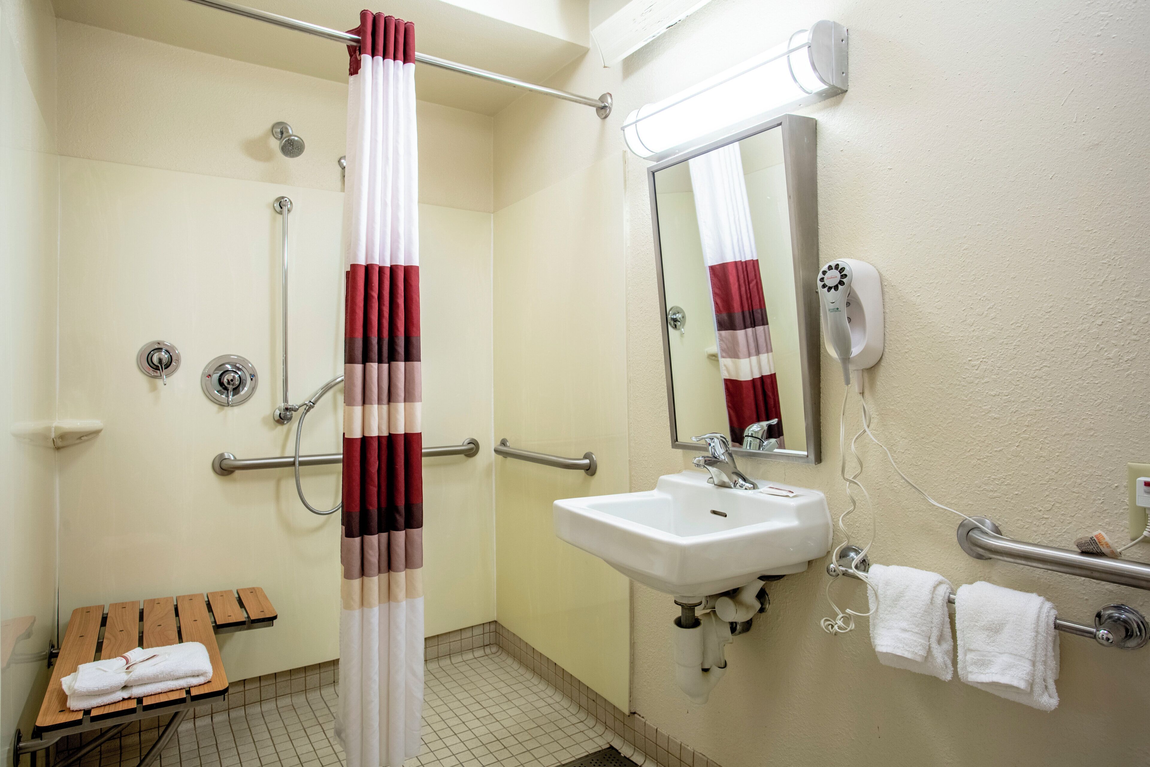 A bathroom with a shower, a sink, a mirror, a towel rack, and a bench with towels.