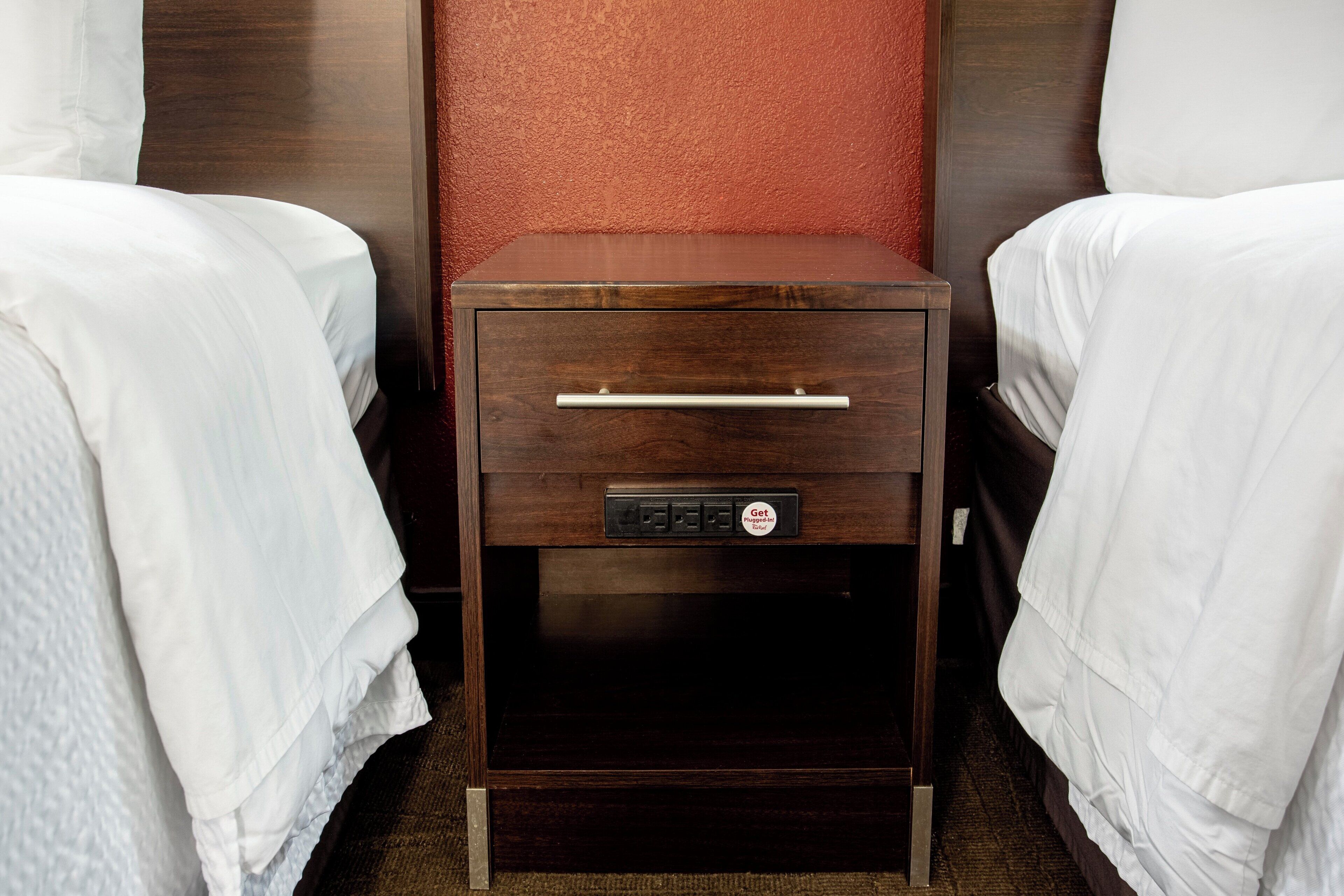 A hotel room with two single beds, a wooden nightstand with a drawer, and a red textured wall.