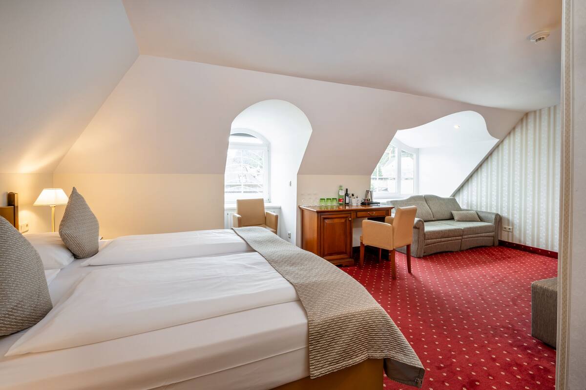 Habitación de hotel con una cama grande, un escritorio, una silla y un sofá. Hay dos ventanas y una alfombra roja.