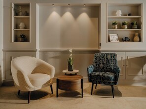 Ein Wohnzimmer mit einem beigefarbenen Sessel, einem gemusterten Sessel, einem kleinen runden Tisch und einem Regal mit Dekorationen.