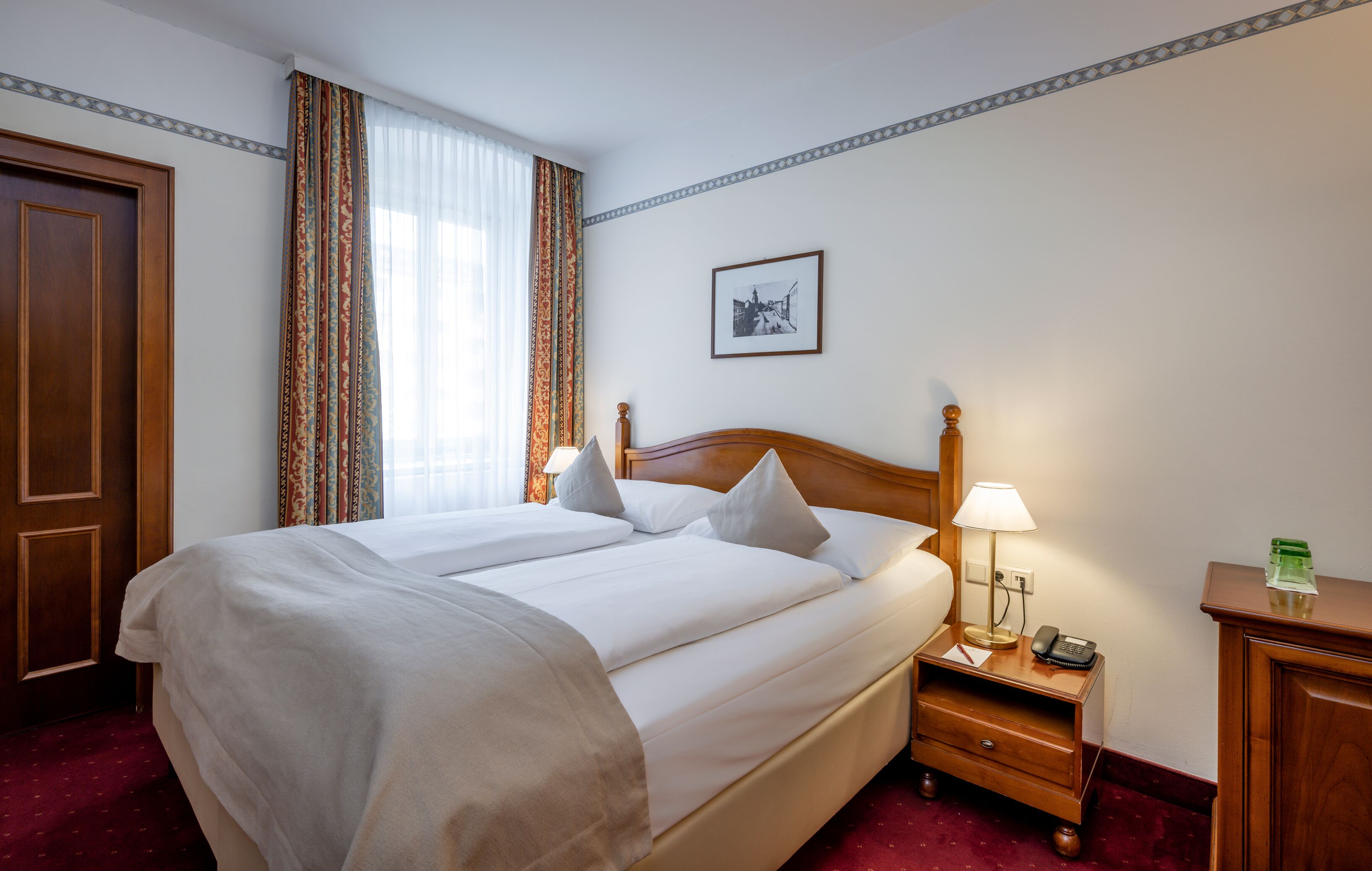 A hotel room with a large bed, a bedside table with a lamp, a phone, and a picture on the wall.