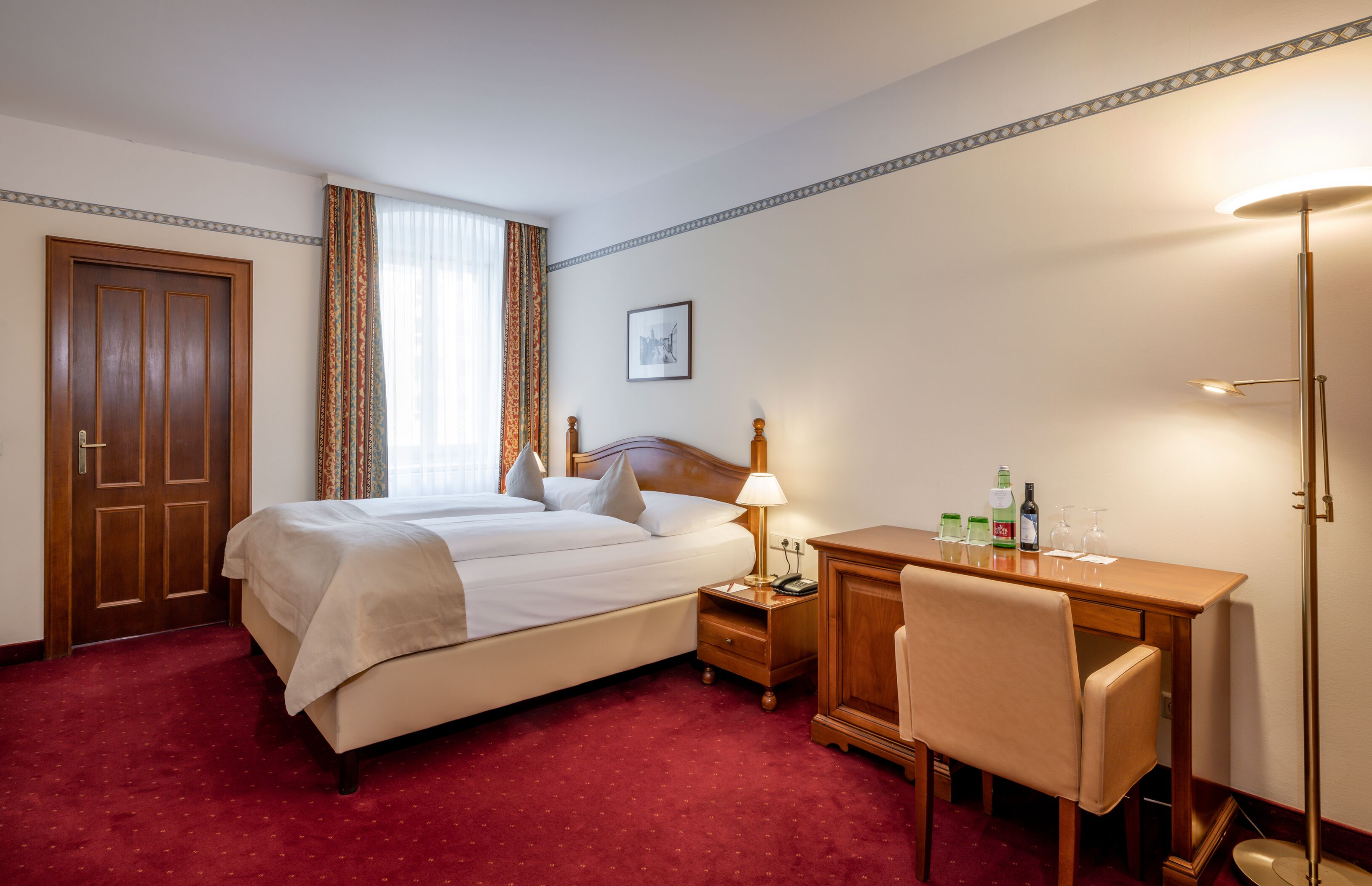 Hotelkamer met een tweepersoonsbed, een bureau met stoel, een nachtkastje en een lamp.