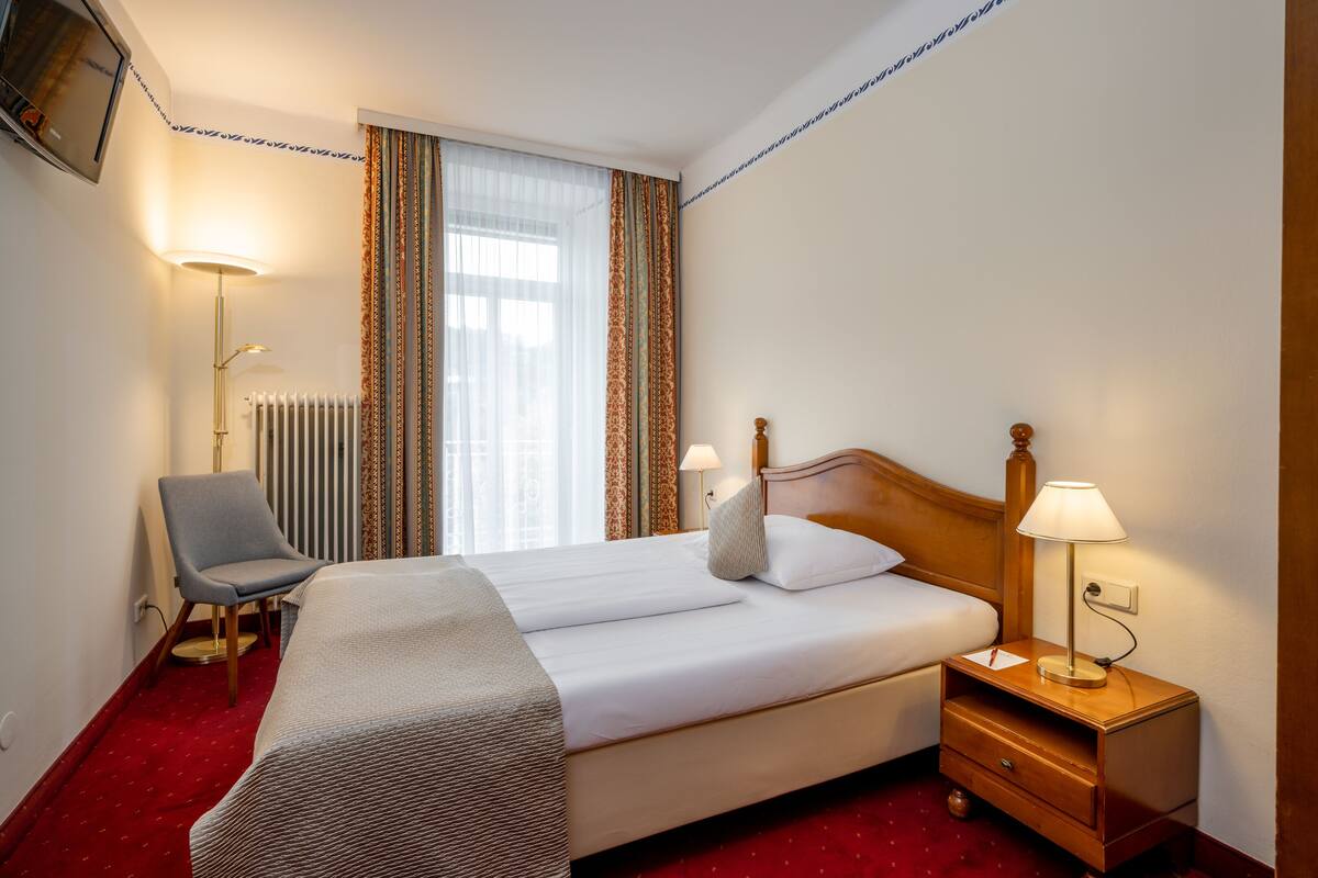 Habitación de hotel con cama doble, una silla, mesita de noche con lámpara, radiador y televisor montado en la pared.