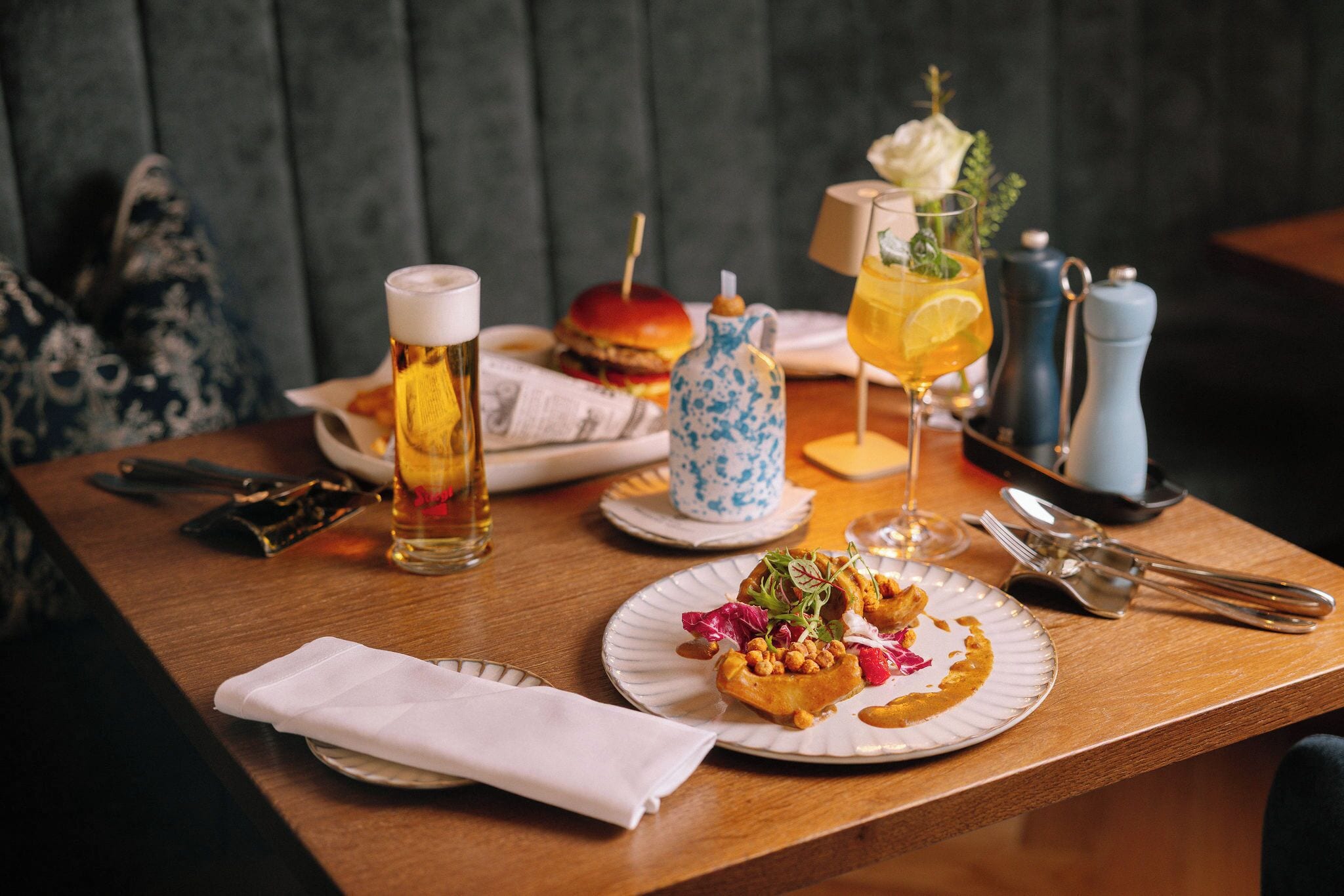 Ein Esstisch mit einem Teller Essen, einem Bierglas, einem Cocktailglas, Salz- und Pfefferstreuern sowie einer kleinen, blau-weißen Keramikkanne.