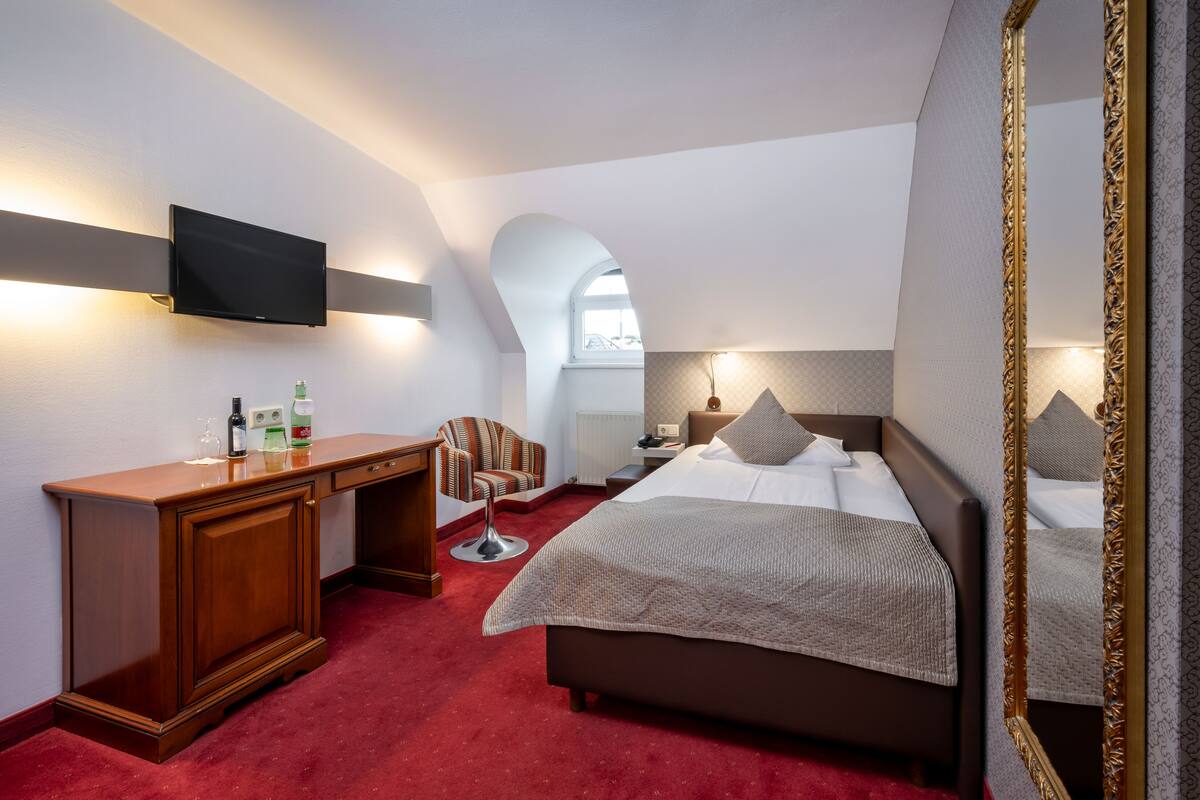 Habitación de hotel con cama, escritorio, silla, televisión y un espejo grande.