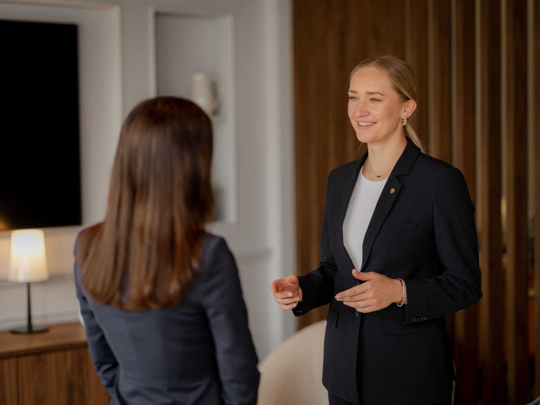 Two individuals in a room with a wooden panel wall, a white door, and a lamp on a side table.