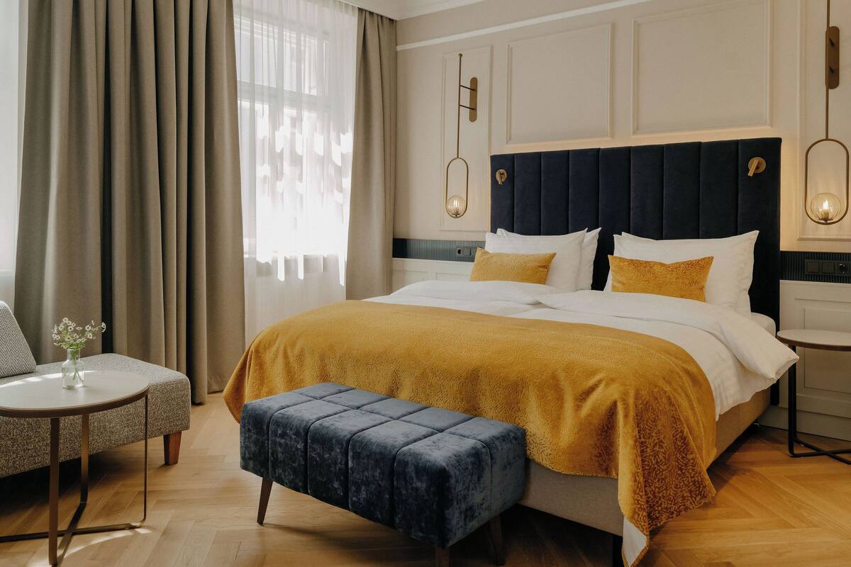 Una habitación de hotel con una cama grande, una mesita redonda, un banco y una ventana con cortinas.