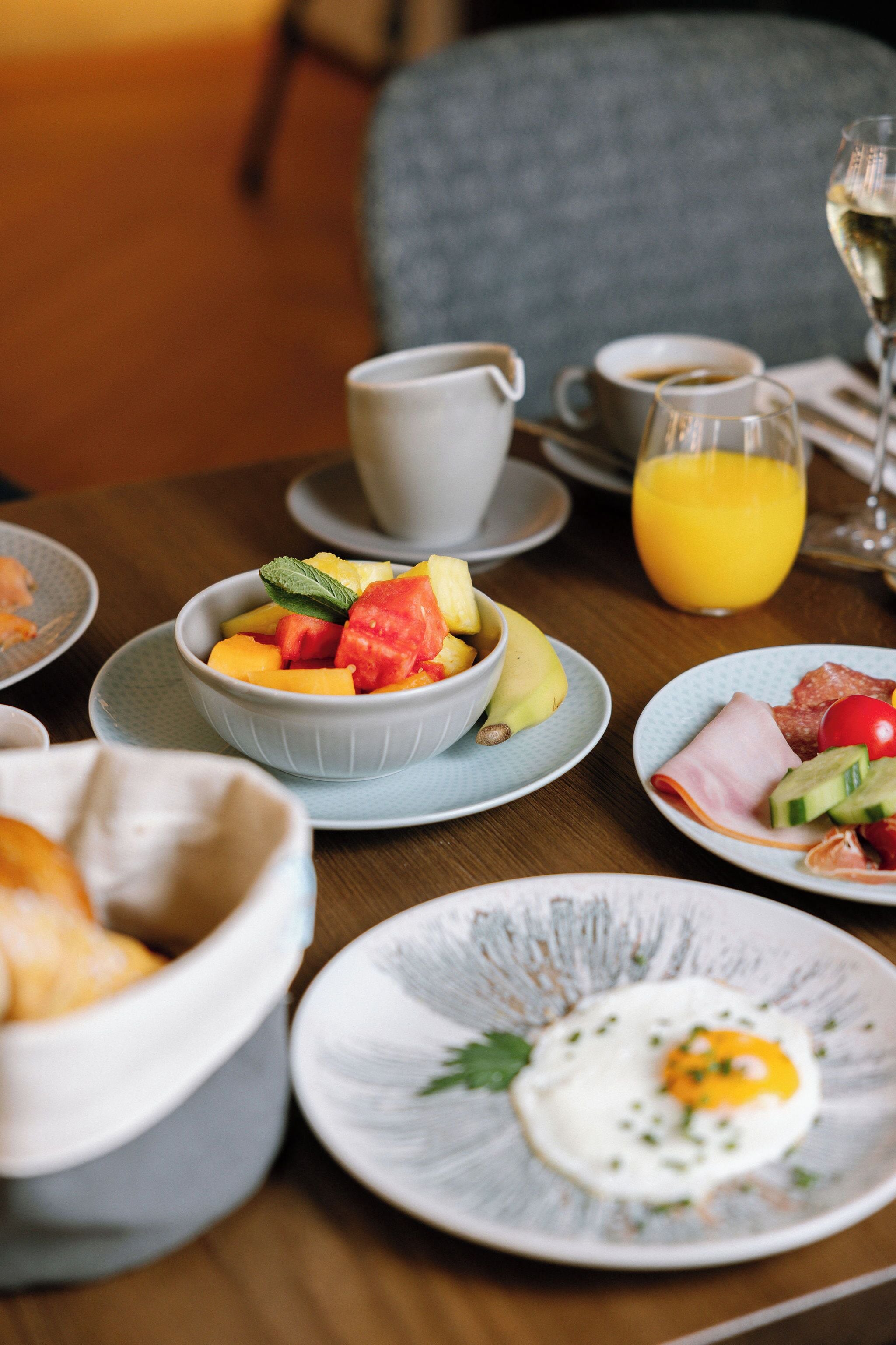 Mesa de café da manhã com uma tigela de frutas, um prato com ovos e um copo de suco de laranja.