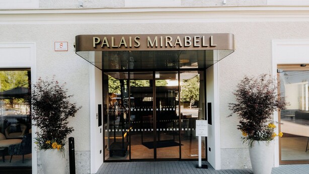 La entrada de un hotel llamado 'Palais Mirabel' con una puerta de vidrio, dos plantas en macetas y un letrero sobre la puerta.