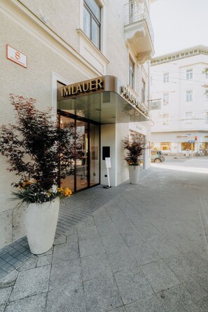 Ein Hotel Eingang mit Glasfassade, einem Schild mit der Aufschrift 'IMLAUER PALAS MIRABEL' und Topfpflanzen auf dem Bürgersteig.