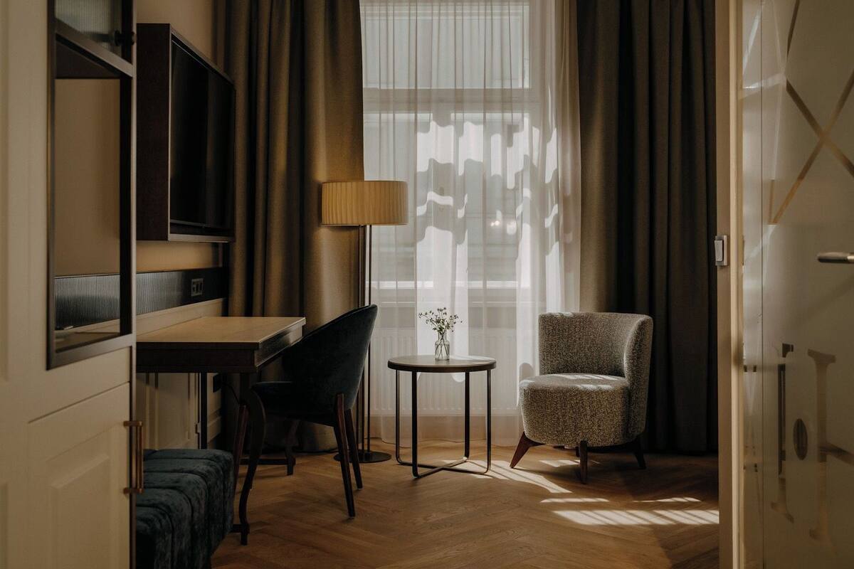 Habitación de hotel con suelo de madera, un escritorio con silla, una pequeña mesa redonda con lámpara, y una cama.