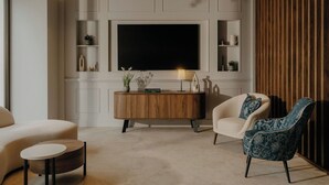 Una sala moderna con un televisor de pantalla plana grande, un mueble bajo de madera, un sofá color beige, un sillón estampado y una pequeña mesa redonda.