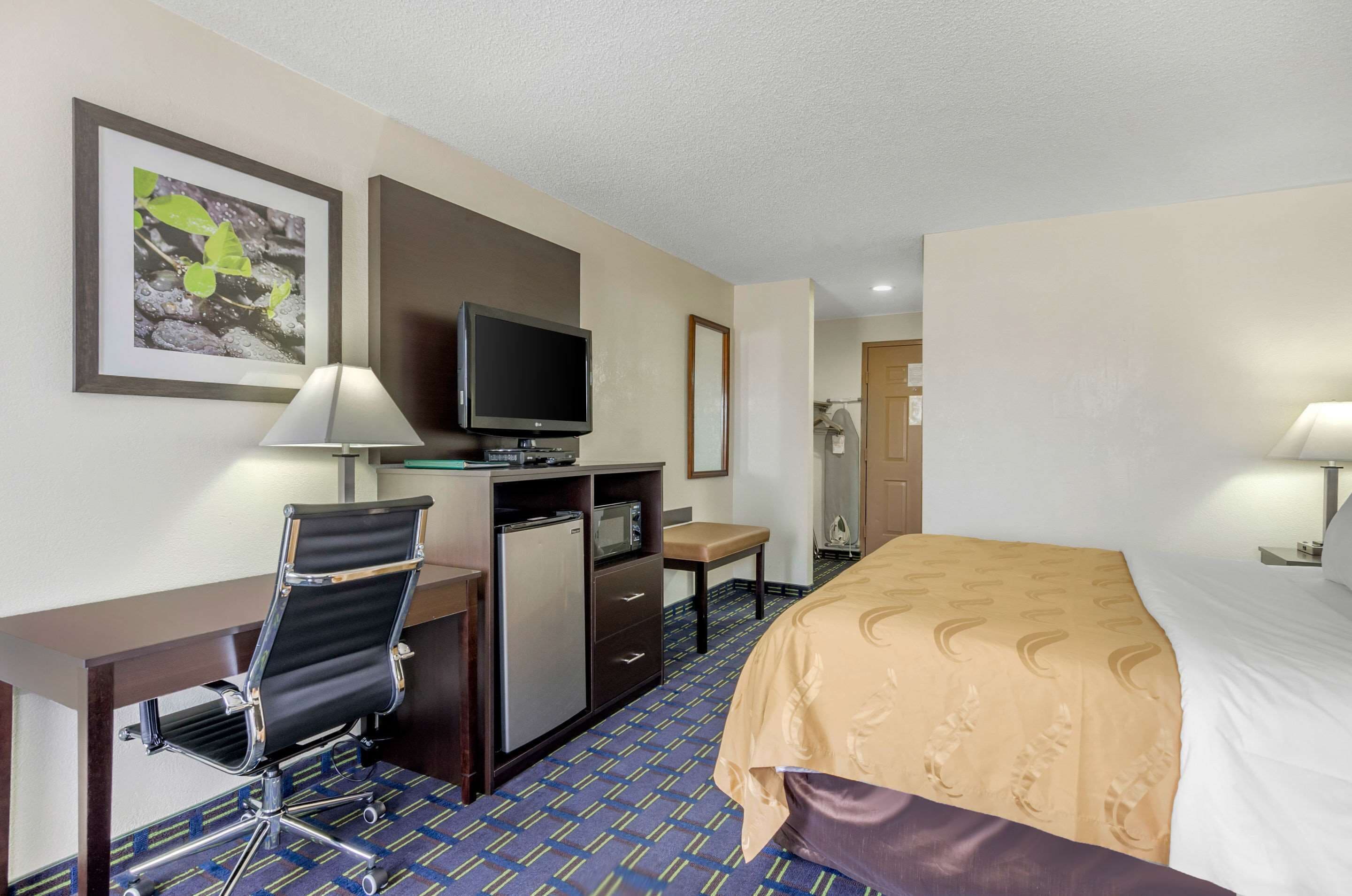 Quarto de hotel com cama, escrivaninha, cadeira, televisão e uma mesinha.