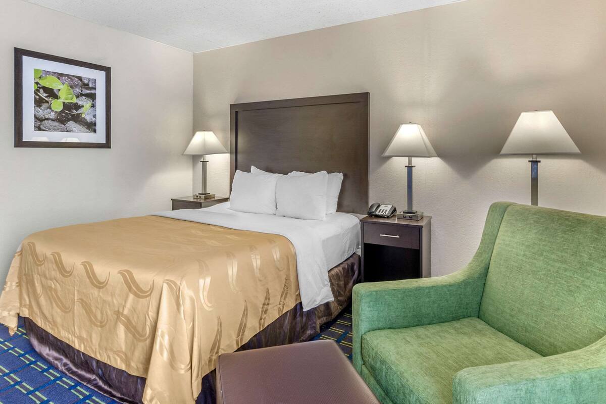 A hotel room with a bed, two bedside lamps, a green armchair, a framed picture on the wall, and a telephone on the nightstand.