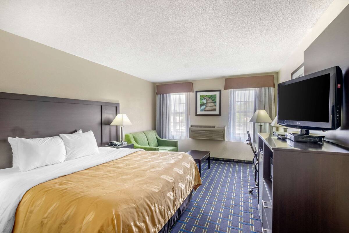 A hotel room with a large bed, a desk with a television, a chair, and a desk lamp.