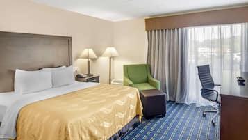 Una habitación de hotel con una cama grande, dos lámparas de noche, un sillón verde, una silla de escritorio negra y un ventanal con cortinas.